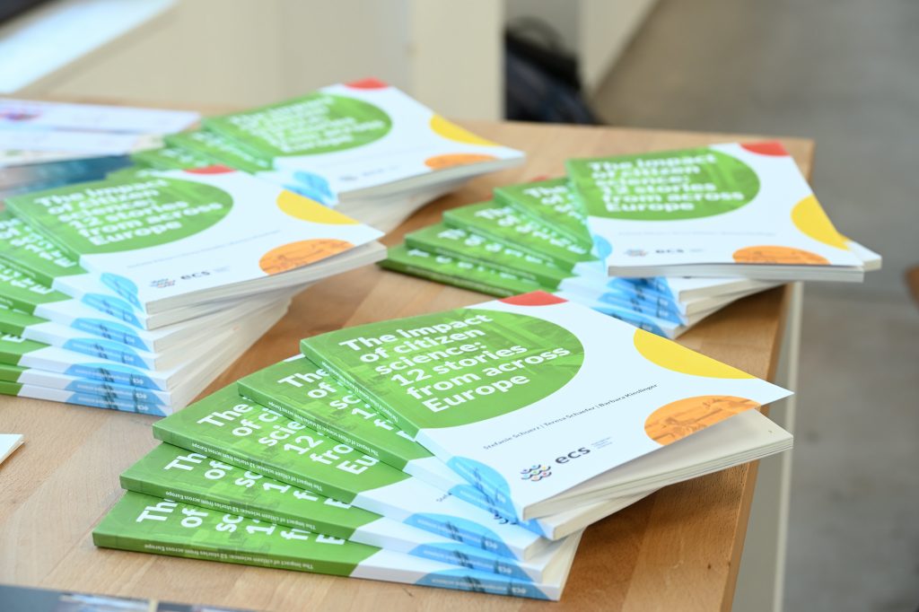 Stacks of books titled The impact of citizen science: 12 stories from across Europe are arranged on a wooden table, ready for distribution at an event hosted by museums and libraries.