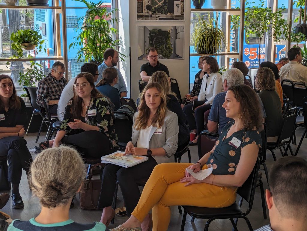 A group of people sits in small discussion circles in a bright room with large windows and many plants. Most participants appear engaged in conversation, holding papers and notebooks.