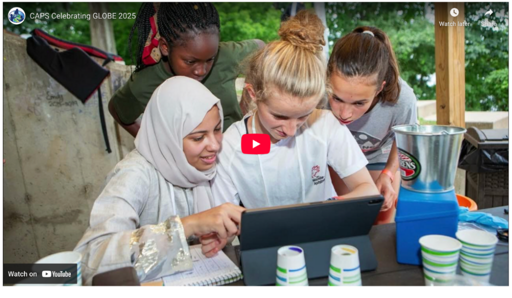 Four diverse students gather around a tablet, smiling and engaged, at an outdoor table with science equipment, paper cups, and a blender visible, suggesting a collaborative STEM activity.