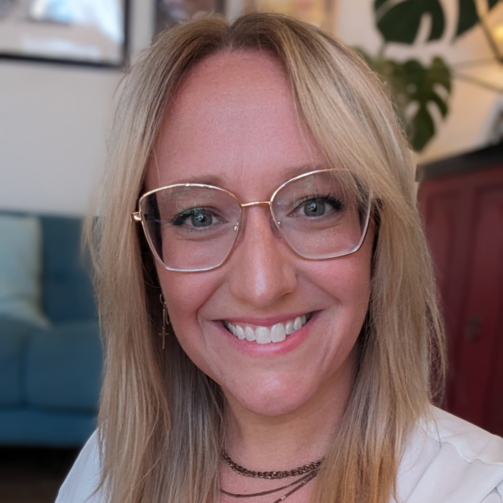 A woman with long blonde hair and large glasses smiles at the camera. She wears a white top, layered necklaces, and a small cross earring. A blue couch and leafy plant are visible in the background.