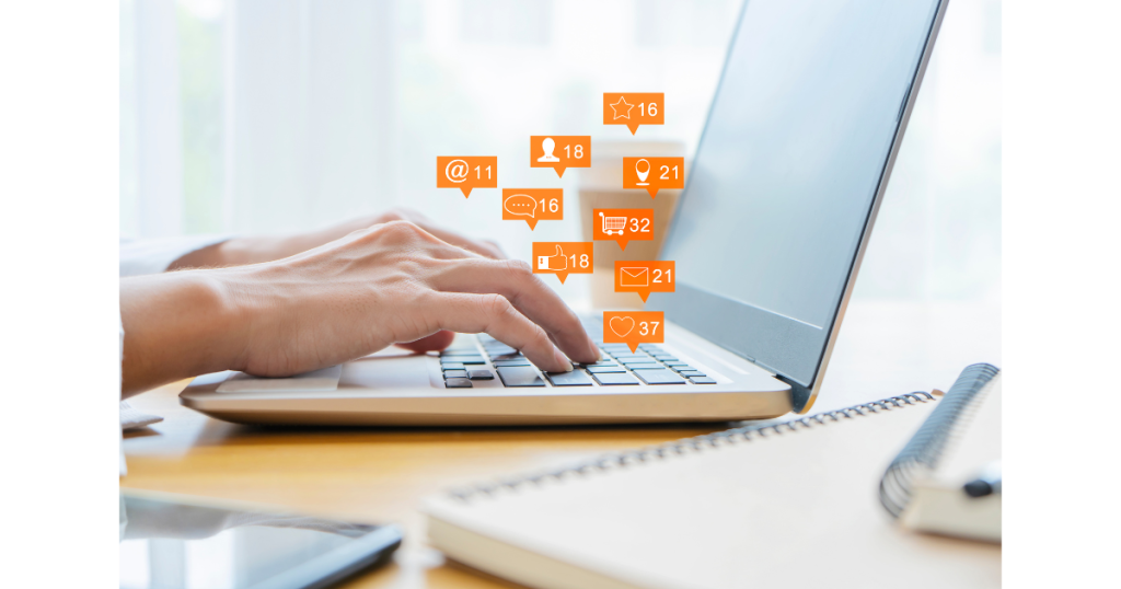 Hands typing on a laptop keyboard with social media notification icons, such as likes, messages, and followers, appearing above the keyboard. A notebook and pen are visible on the desk.