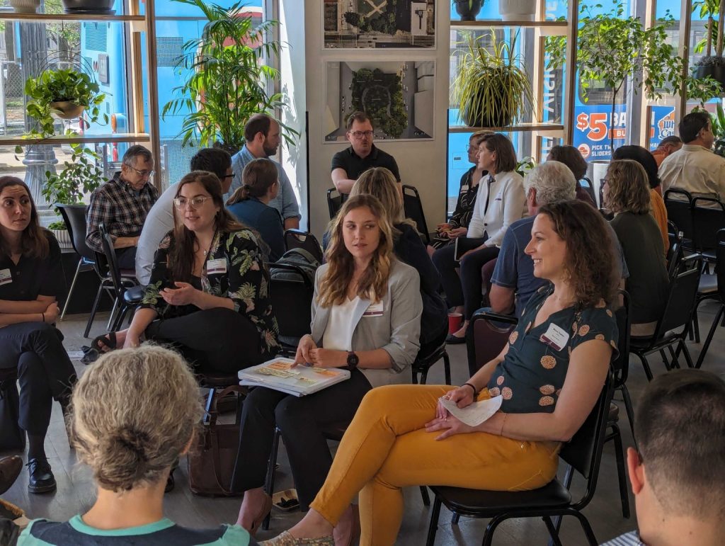 A group of adults sit in small circles, engaged in conversation in a sunlit room filled with plants and large windows. Some people hold notebooks and wear nametags.